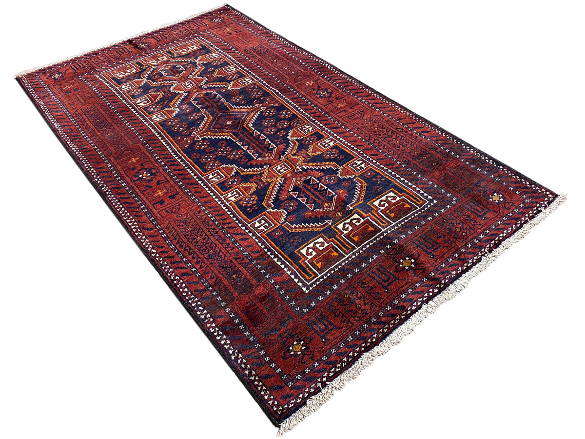 Tapis baloutche rectangulaire, rouge, avec un champ ornemental dense en bleu/blanc et une bordure périphérique.