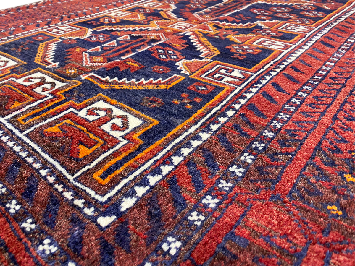 Gros plan d’un tapis baloutche : motif rouge et bleu avec des lignes décoratives blanches et jaunes.