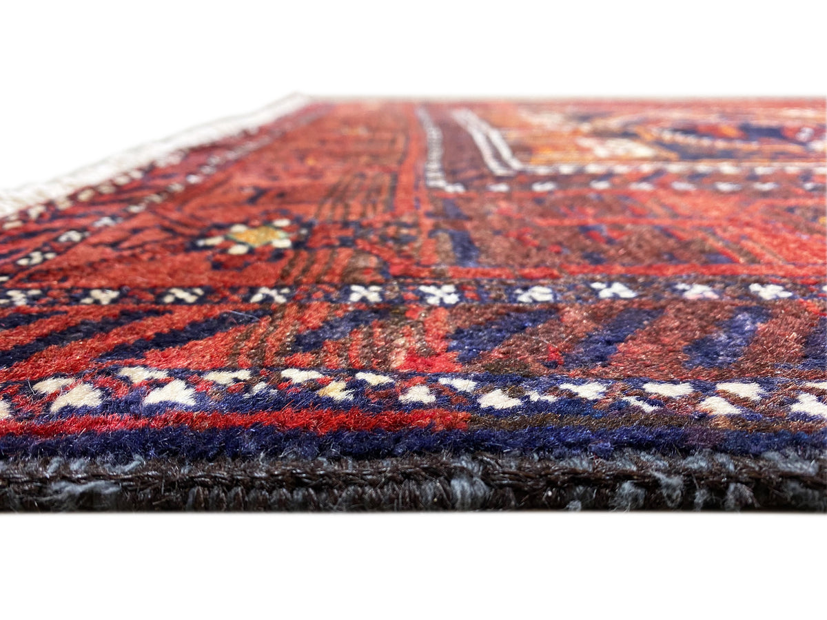 Détail de bordure d’un tapis baloutche, champ ornemental rouge et bleu et franges foncées sur le bord.