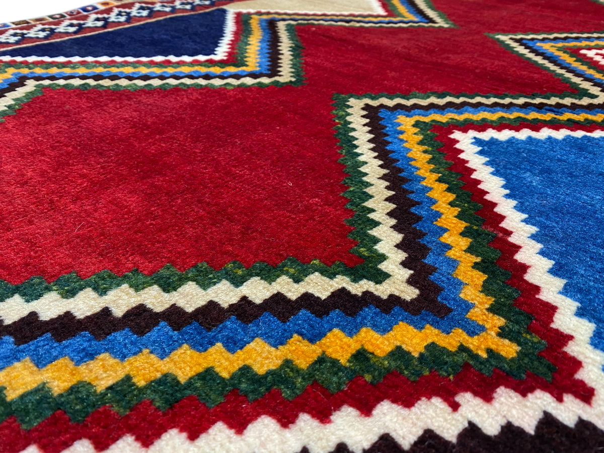 Nahaufnahme: Rotes Mittelmotiv und farbige Zickzackkanten eines Shiraz-Teppichs, mehrfarbige Bordüre.